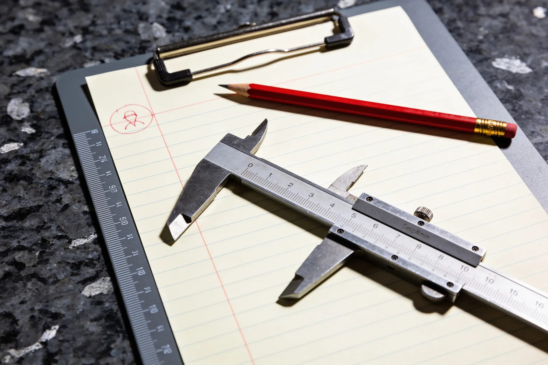 A blank machinist's inspection sheet clipped to a board on a granite surface plate, with a steel caliper and a red pencil laid across it.